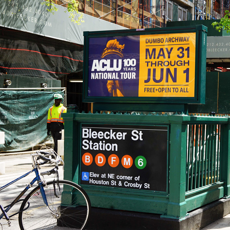 A subway station in New York City with an ACLU tour ad. The ad is split into two segments. On the left is a duotone image of the statue of liberty torch in yellow on a navy blue background, with the "ACLU 100 Years National Tour" logo across it. On the right is the event details, in navy blue on a yellow background.