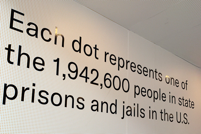 Another view of the same exhibit. Large text reads "Each dot represents one of the 1,942,600 people in state prisons and jails in the U.S." The wall and ceiling visible in the photo is covered in a grid of tiny orange dots.