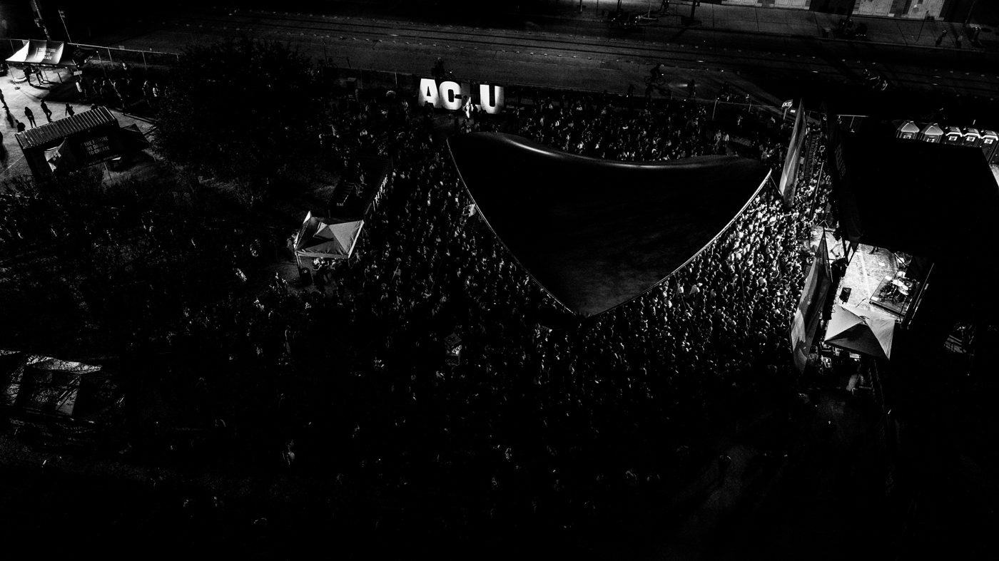 A black-and-white photograph of a crowded event. There is a stage at the right side of the photo and a large audience. At the edge of the audience is a large sign with the letters ACLU.